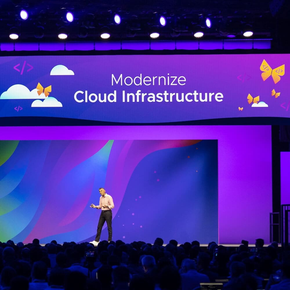 A person is hosting a large conference talk in front of a screen that reads "Modernize Cloud Infrastructure."