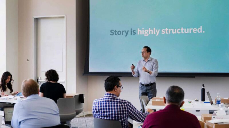 A person leads a presentation in front of a classroom, with "Story is highly structured" on the screen behind them.