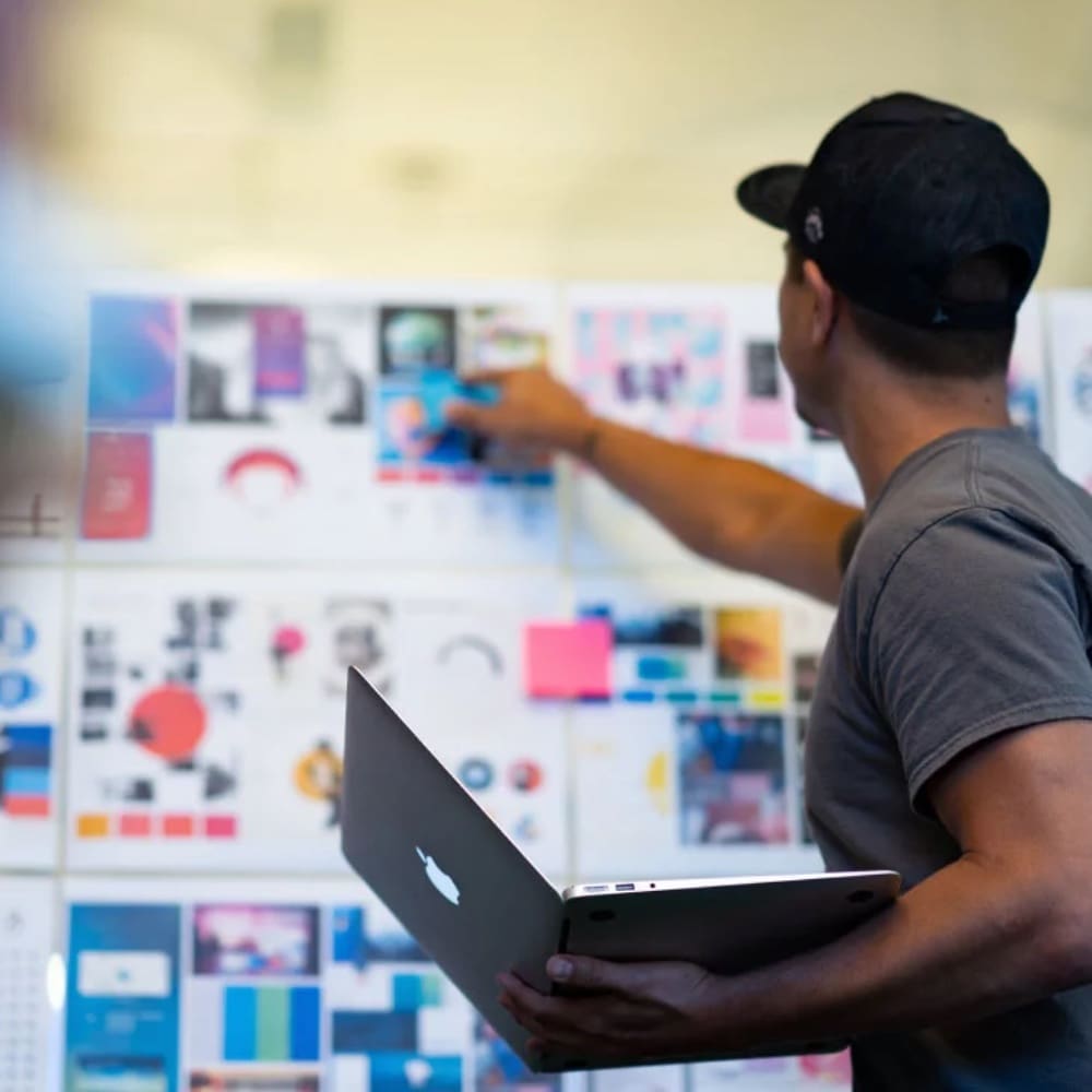 A Black man holding a laptop points at a vision board with slides, images, and sticky notes.