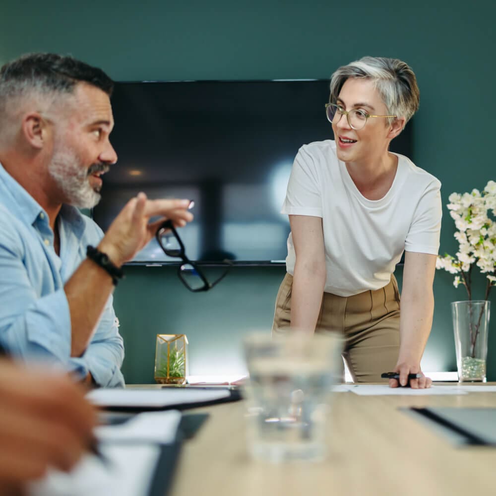 A person standing at a conference room table is discussing something with a man sitting next to them.