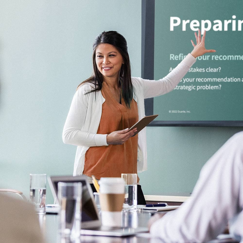 A woman points at a screen and leads a presentation in front of a conference room.