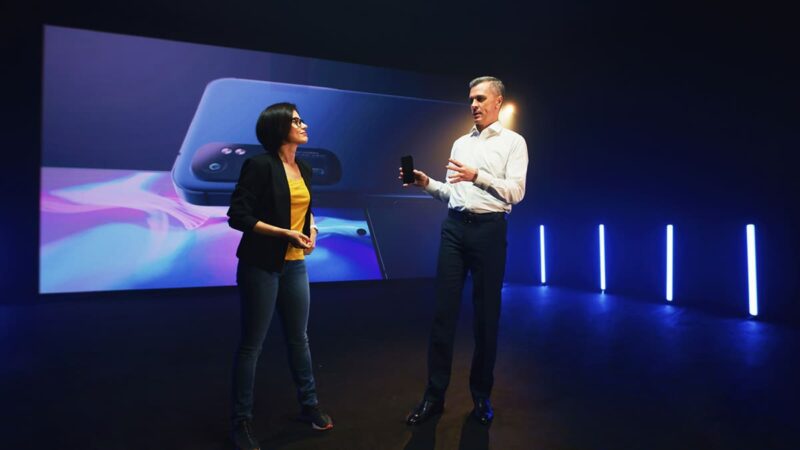 Two people on a darkly lit stage discuss a smartphone with a closeup image of the device behind them.
