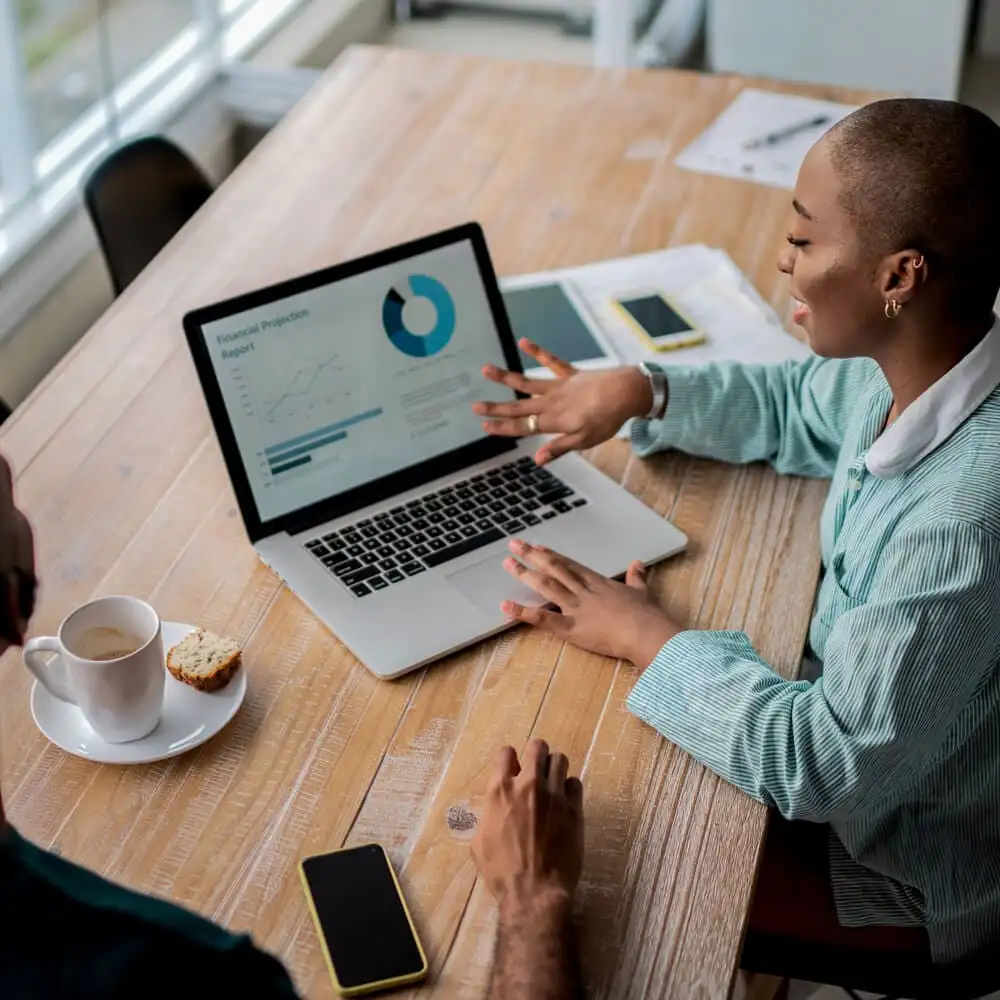 A Black person points at their laptop while discussing a presentation slide with another person.