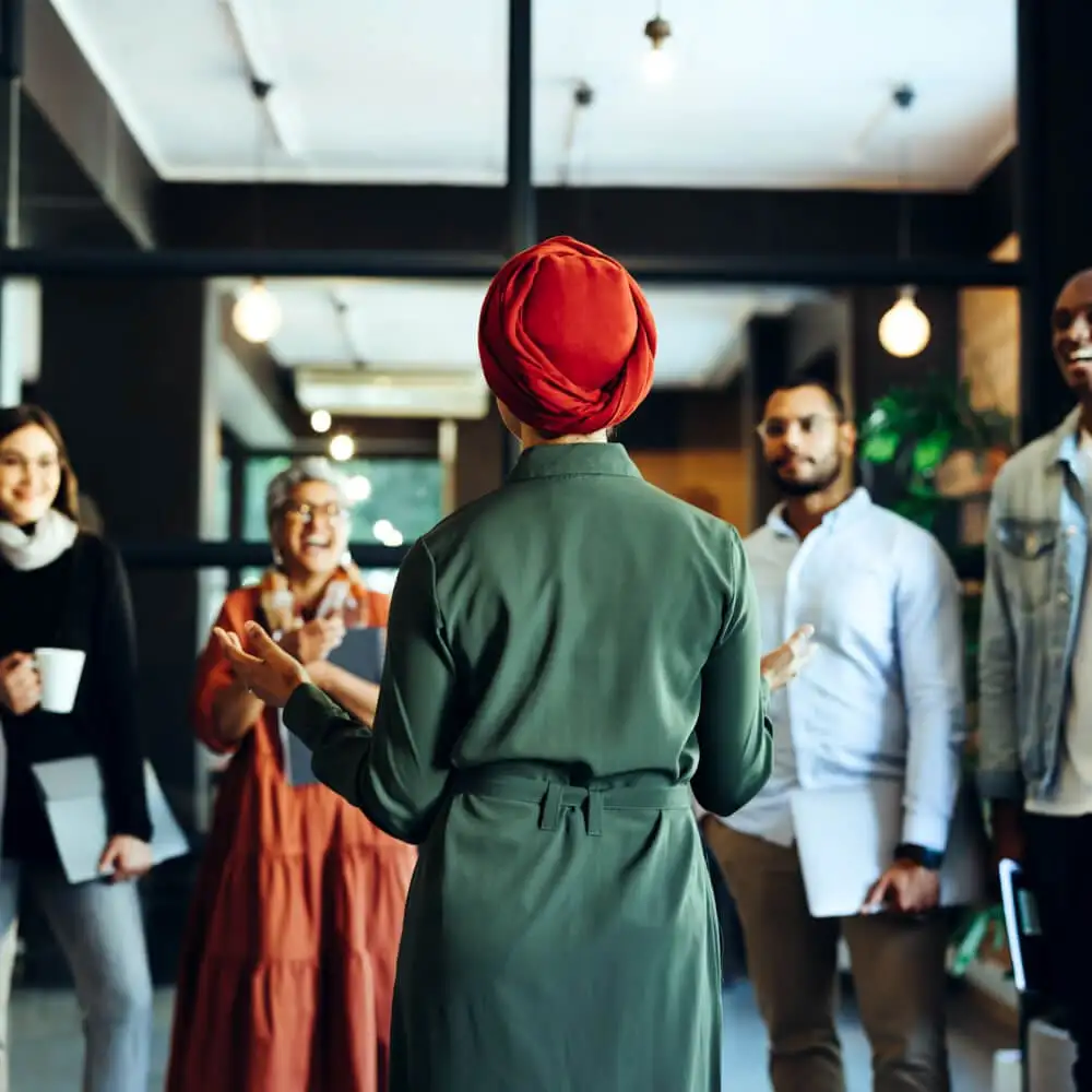 A person with a head wrap talks in front of a group of people while everyone is standing.