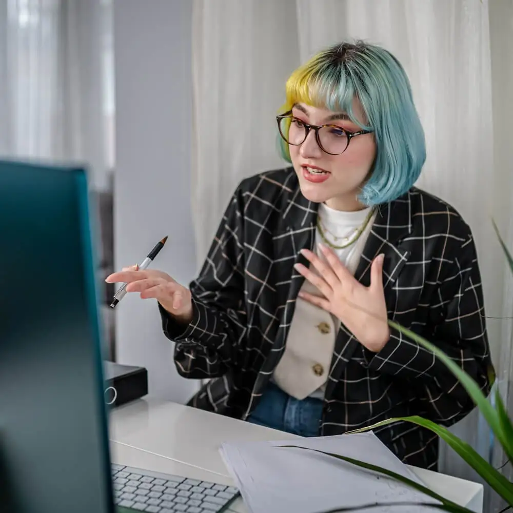 A woman with colorful hair talks during a virtual meeting on her computer.